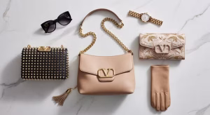 Composición flat lay de tres bolsos icónicos Valentino Garavani sobre mármol: Rockstud negro, VSLING beige y clutch DeVain bordado con accesorios de lujo