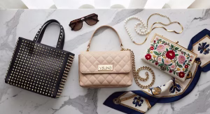 Composición flat lay de tres bolsos icónicos Valentino Garavani sobre mármol: Rockstud negro, VSLING beige y clutch DeVain bordado con accesorios de lujo